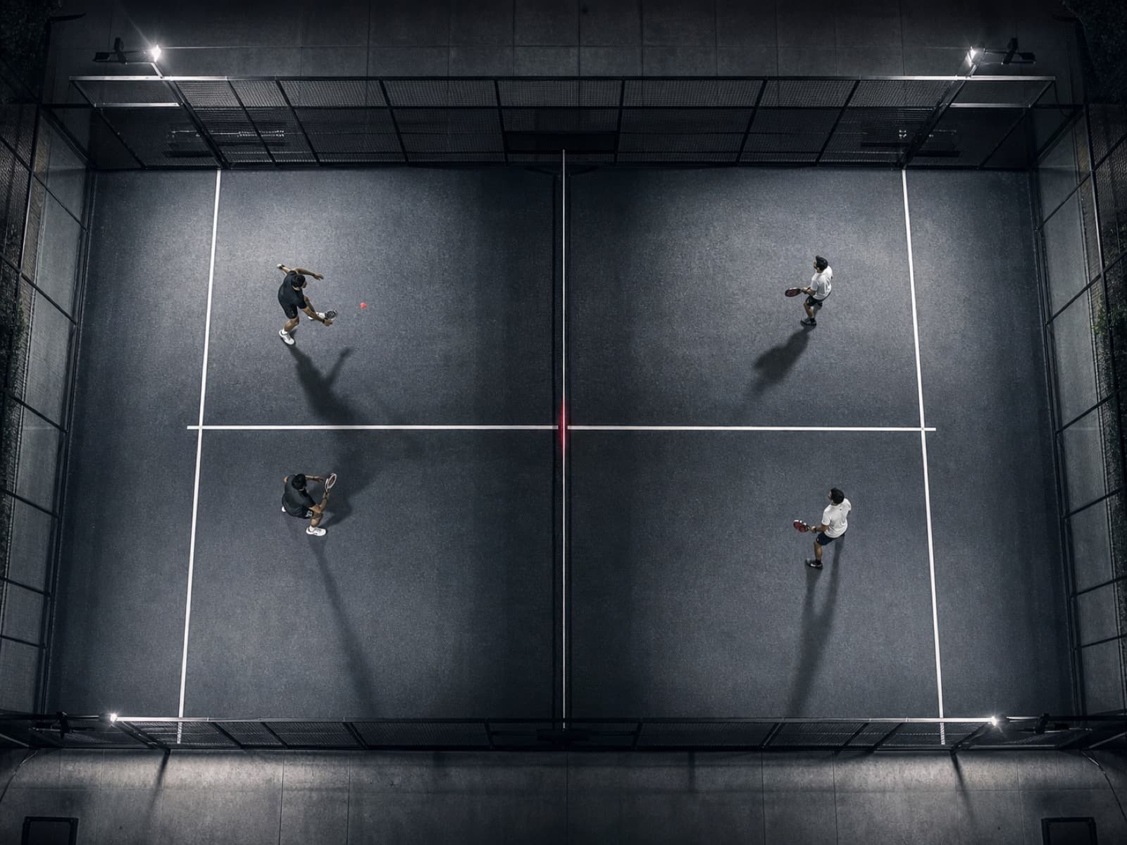 Overhead view of a floodlit padel court at night with four players mid-rally